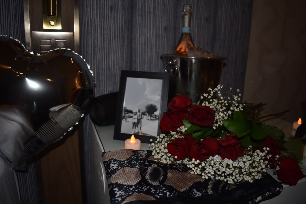 A romantic setup featuring a heart-shaped balloon, a framed black and white photo, a bouquet of red roses with baby's breath, a silver champagne bucket with ice, and a candle, all arranged on a table.