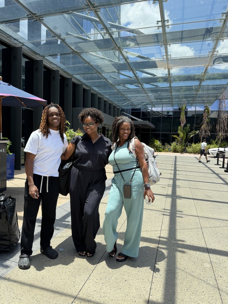 Three individuals standing together in a modern outdoor space, smiling and posing for the camera, with a transparent glass roof above and greenery in the background.