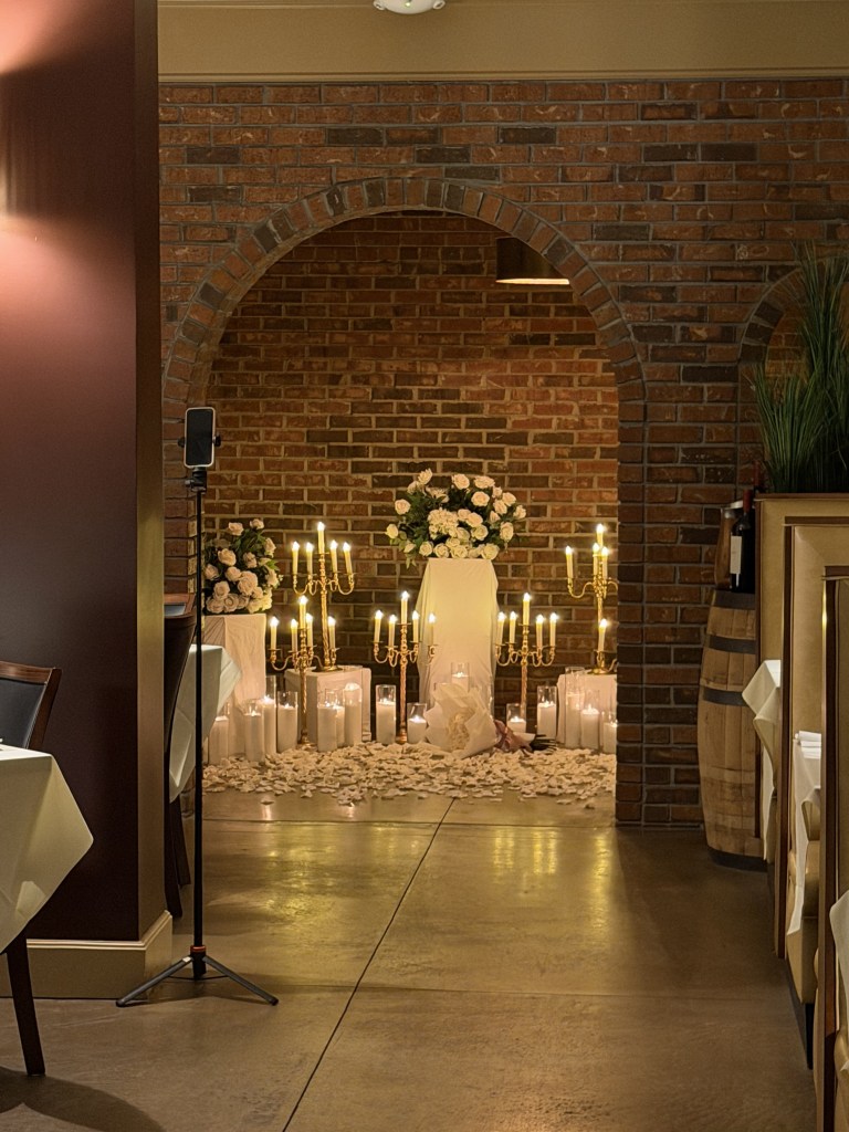 A beautifully decorated indoor space featuring a brick wall archway. In the foreground, there are elegant white tablecloths on tables and a microphone stand. The background showcases a romantic arrangement of candles, white flowers, and rose petals, creating a warm ambiance for a special event.