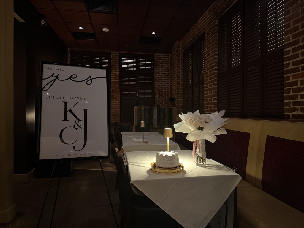 A decorated table in a restaurant featuring a sign that says 'She said yes! Let's celebrate! K & J', a small cake on a gold stand, and a floral arrangement in a vase.