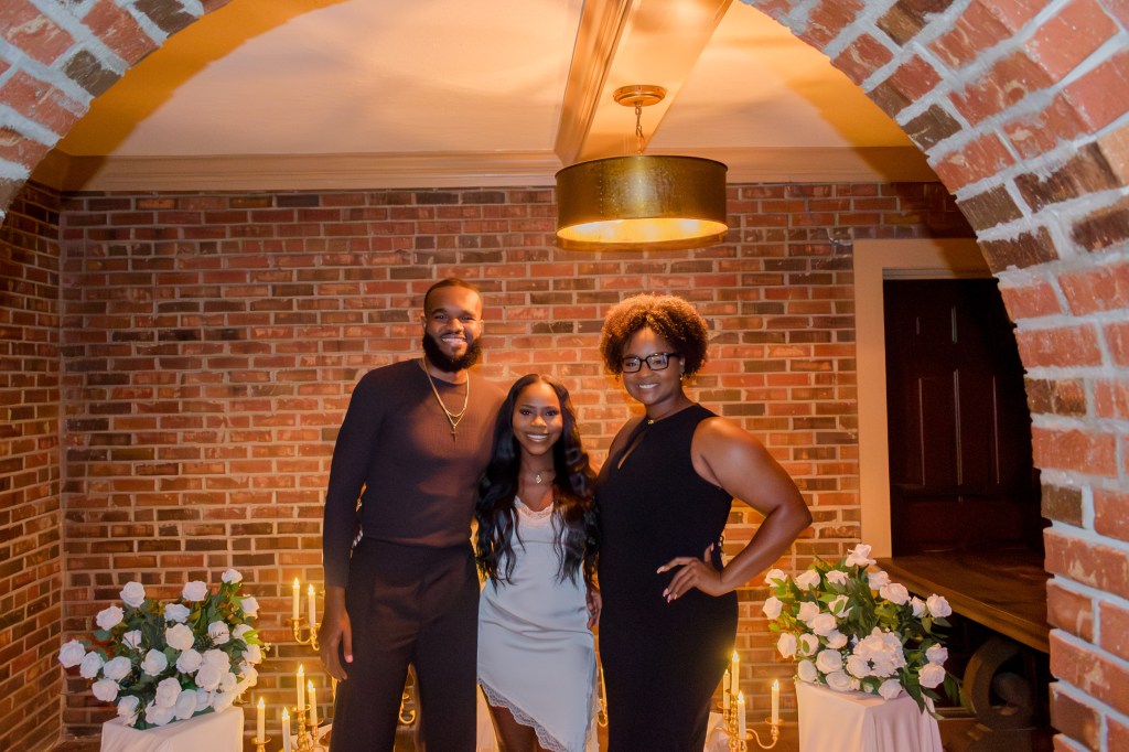 A group of three people posing together in a warmly lit indoor setting, with brick walls and decorative flowers on display.