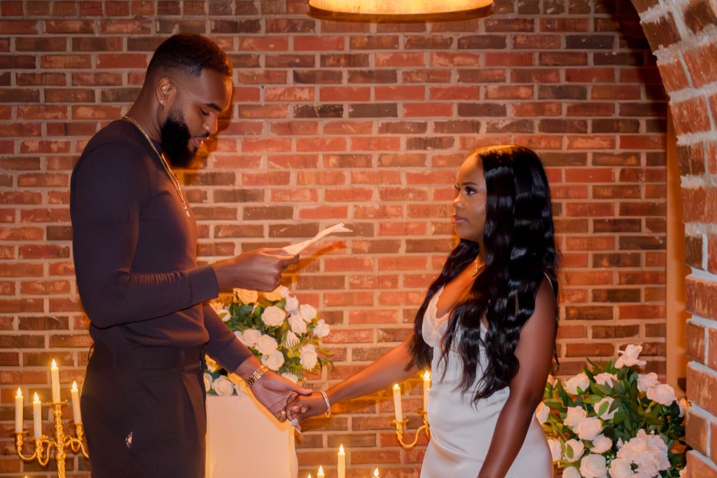 A man in a fitted black outfit reads a note while holding hands with a woman in a white dress. They are in a dimly lit space with brick walls, candles, and flowers in the background.